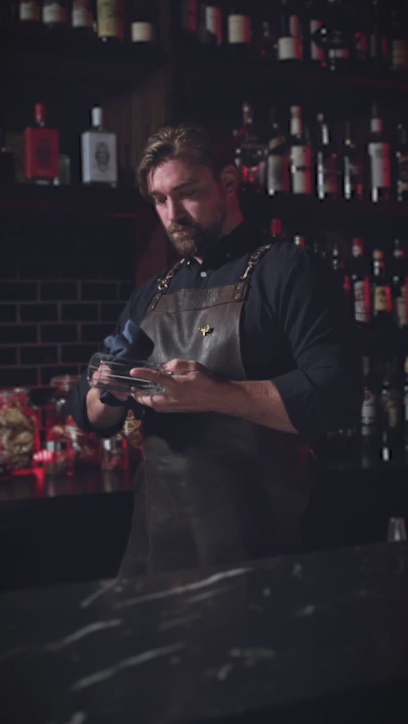 Man in an apron working behind a bar with bottles on shelves in the background.