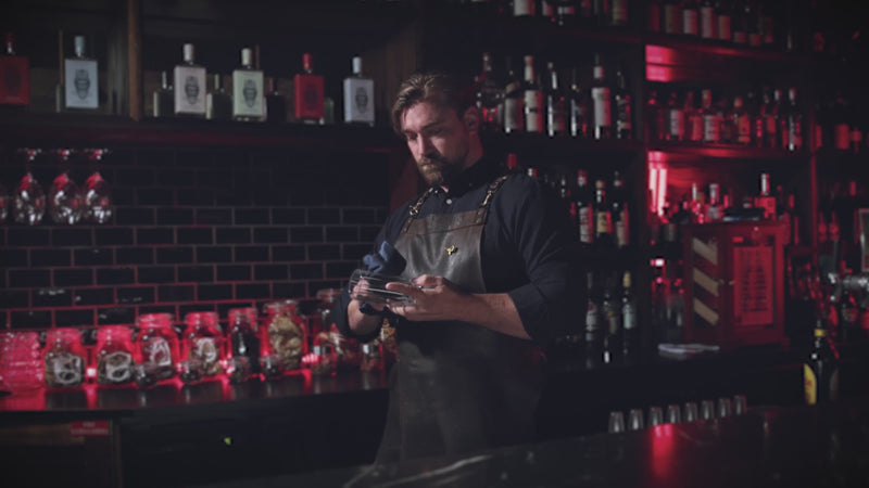 Bearded bartender in black attire and apron mixing a drink in a cozy bar.